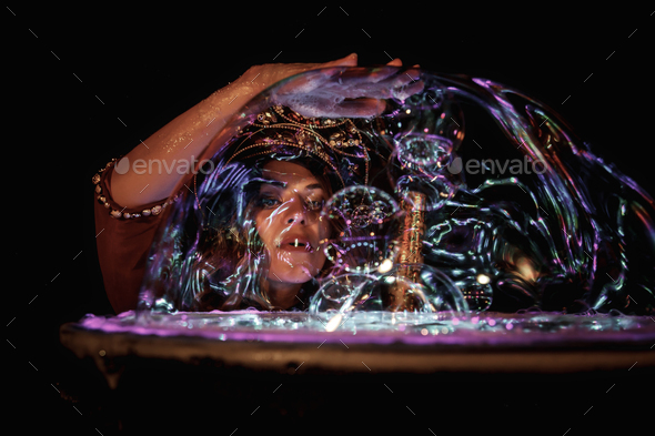 Female magician illusionist of circus showing soap bubbles show on tabletop at black background