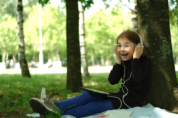 Little girl in headphones sitting in the park with her digital tablet listening to music or lessons