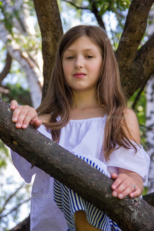 A little Caucasian girl with blonde hair and a white blouse is sitting on a branching tree. It\'s sum