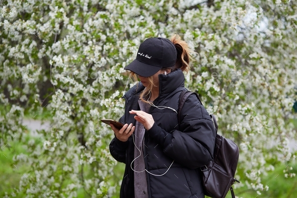 Stylish girl surfing in phone, listen to music outside near blooming tree. Spring walking