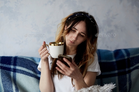 Young aesthetic woman using phone and drinking tea at home. Cozy home, freelance, leisure time.