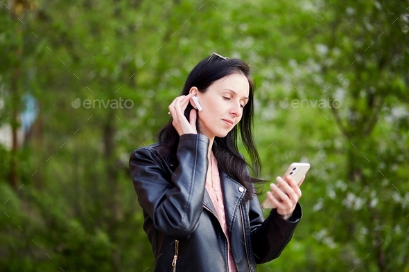 Young stylish woman listening music, talking using headphones and mobile phone in the park