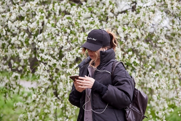 Stylish girl surfing in phone, listen to music outside while walking among spring park