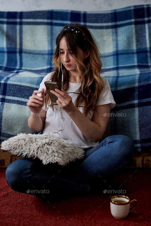 Girl sitting on the floor, using phone, listening to music and having a tea at cozy home