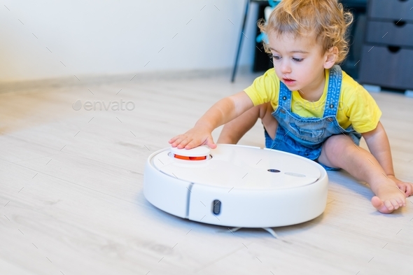 little boy 2 years ago in a yellow T-shirt and denim overalls turns on a white robot vacuum cleaner