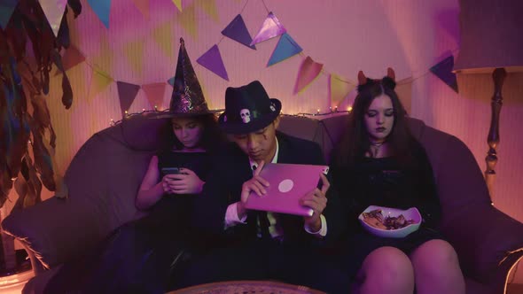 A Guy in a Creepy Costume is Actively Playing on a Tablet and the Girls are Sitting on the Phone at alt