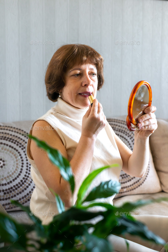 A beautiful woman 45-50 years old paints her lips with lipstick looks in the mirror. Self care