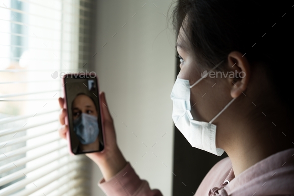 Portrait of a young girl wearing a disposable medical mask, using a  mobile phone for a video call - Stock Photo - Images