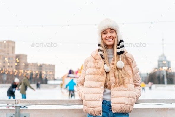 Woman in fluffy fur hat winter jacket standing outdoor near ice skating rink. Christmas mood