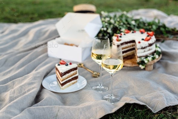 Two glasses of white wine and a cake on a picnic blanket during sunset time outside.