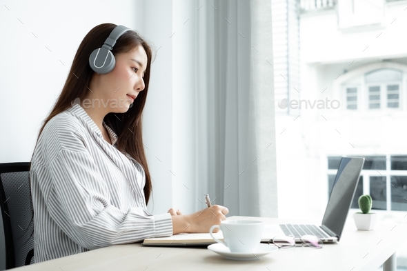 female Asian college student wearing headphones enjoys online tutoring, watching videos and taking n