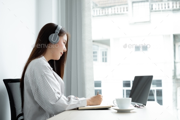 female Asian college student wearing headphones enjoys online tutoring, watching videos and taking n