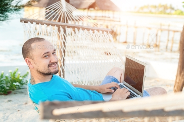 A man works on a laptop while lying in a hammock on the beach.
