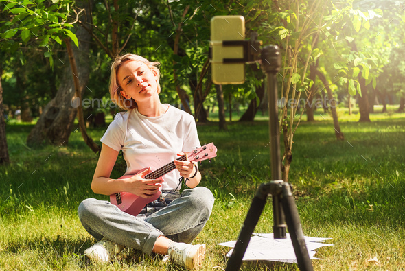 Beautiful teen girl playing ukulele in front of phone on tripod