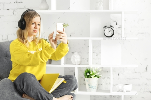 Girl in earphones in yellow blouse sit relax on grey couch use smartphone to listen music, courses.