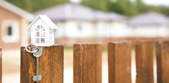 Small figure of white wooden house on fence with key to housing against the background of cottages.  - Stock Photo - Images