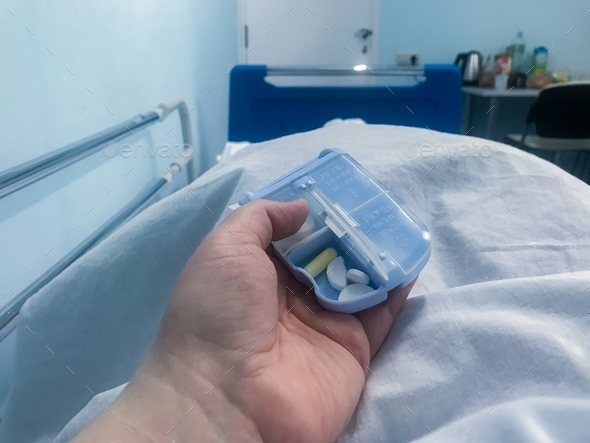 Lying in a hospital bed, holding an open pillbox with pills white and yellow. In a hospital room