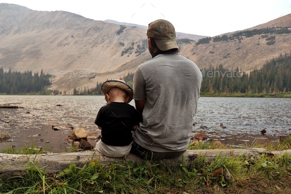 A father and son enjoying the mountain views together while sitting on a log at the lake.  - Stock Photo - Images