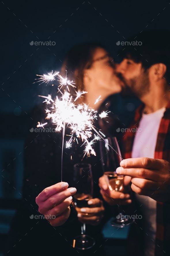 Young woman and man kissing while holding champagne flutes and sparkles