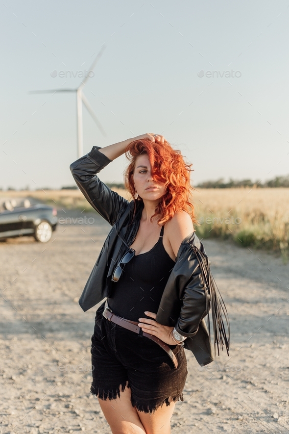 Young woman with red hair in a black leather jacket and sunglasses in a field with windmills