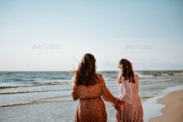 Two girls with long hair from behind holding hands wearing dresses at summer beach in golden hour