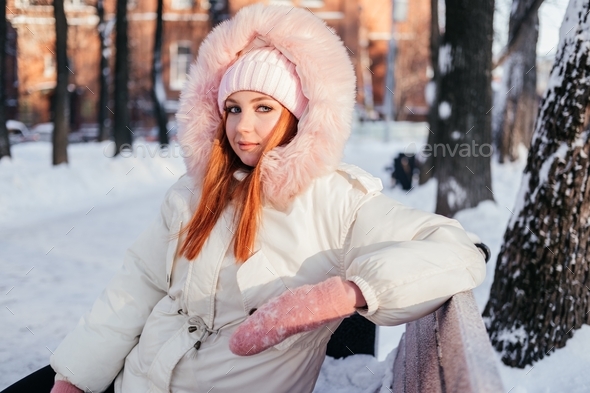 Ginger young woman in coat, hat, mittens sitting on bench in winter park outdoors
