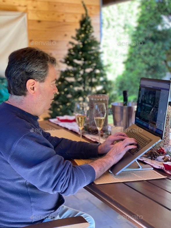 man sitting in the back patio with champagne on a zoom call with a christmas tree and decorations
