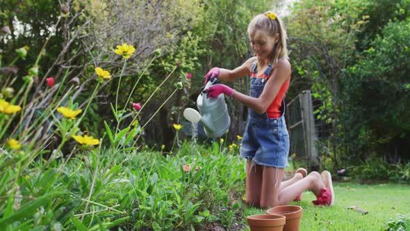 Happy caucasian girl in garden kneeling and watering plants with watering can alt
