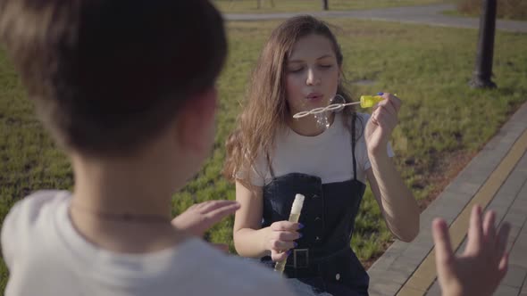 Portrait of the Little Boy Looking at His Older Sister in the Park Who Blowing Soap Bubbles alt