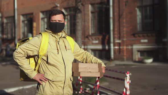 Delivery man in black mask with yellow backpack and pizza boxes stands on street