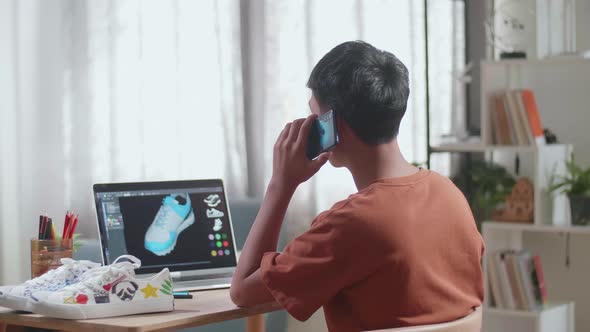 Asian Boy Footwear Designer Talking On Smartphone While Designing Shoe On A Laptop At Home alt