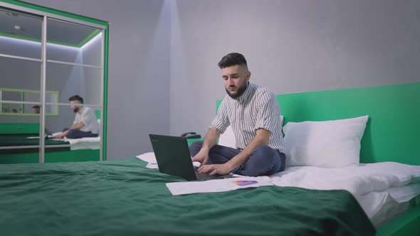 Wide Shot of Busy Young Handsome Businessman Analyzing Documents and Emarket Sitting on Bed in Green alt
