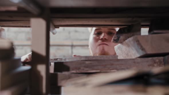 Bearded Man in a Carpentry Workshop Takes a Wooden Detail From the Shelf and Blowing Dust From It alt
