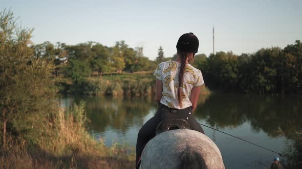 A young girl sits down to the lake while sitting on a horse. Rider in a helmet for riding. Jockey. alt