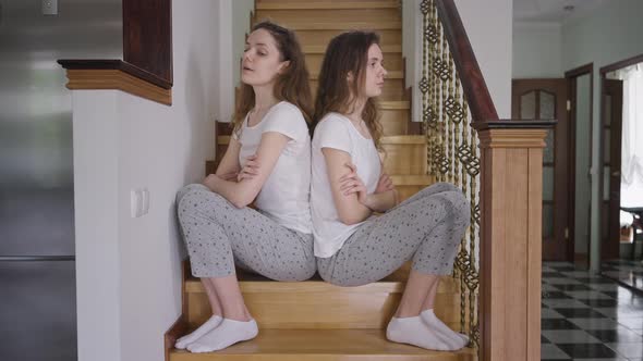 Dissatisfied Argued Twin Sisters in Pajamas Sitting on Stairs Back to Back alt
