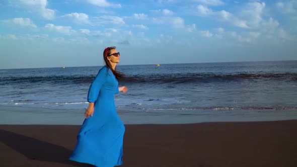 Woman in a Blue Dress Runs Along a Black Volcanic Beach alt