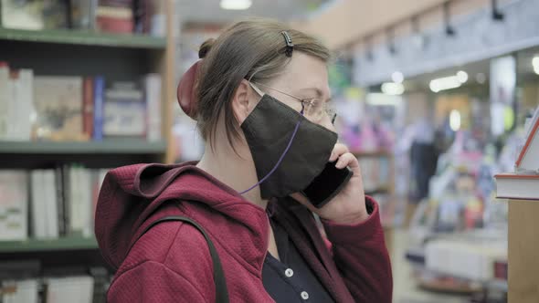 Woman in an Against Virus Mask and Glasses Stands in Shop and Talks on Phone