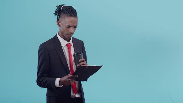 Black Male Businessman in Jacket Red Tie and Dreadlocks Stares Intently Into Paper Tablet and Takes alt