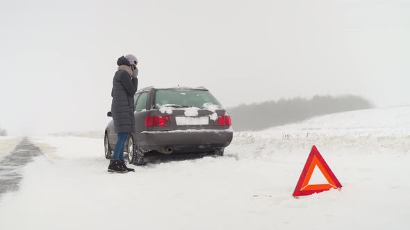 Woman Walks Around Brokendown Car and Calls on Her Smartphone alt