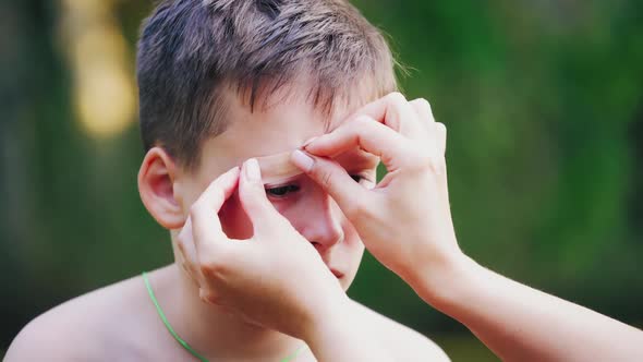 Sad boy with injury on his eyebrow. Woman's hands putting small white adhesive plaster alt