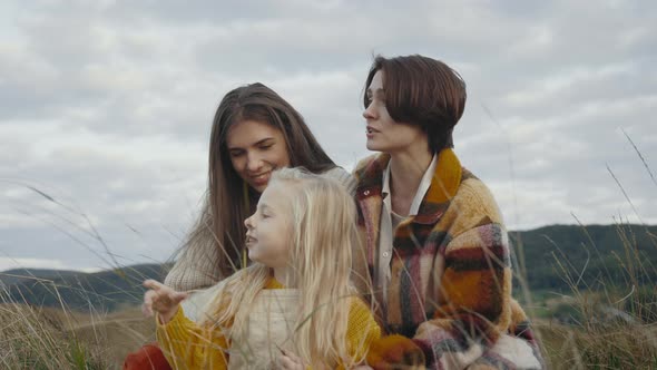 Happy Female Parents Sitting in Grass with Pretty Daughter alt
