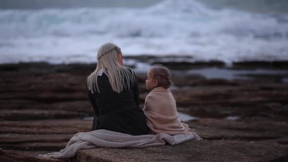 Back View of Blond Woman with Little Girl Cuddling in Plaid on Rocky Shoreline in Twilight Time