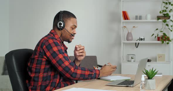 Side View African Student Guy Listen Tutor Through Wireless Headset Writes Information in Notebook alt