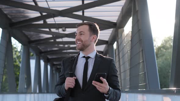 A Young Man in Business Clothes Walks Across the Bridge and Smiles alt