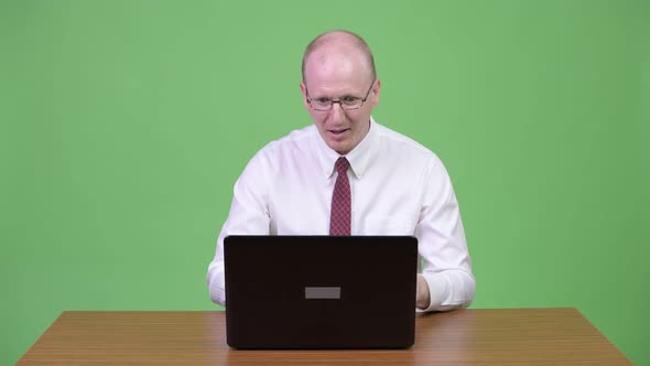 Happy Mature Bald Businessman Using Laptop and Looking Surprised Against Wooden Table alt