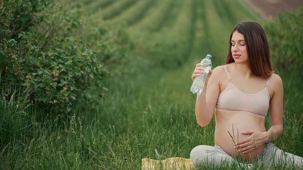 Pregnant young woman with dark hair in a pink top and light pants drinks water outdoors.