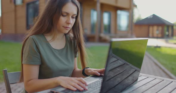 Thoughtful Serious Girl Typing Text on Laptop Keyboard Sitting at Table in Yard of House alt