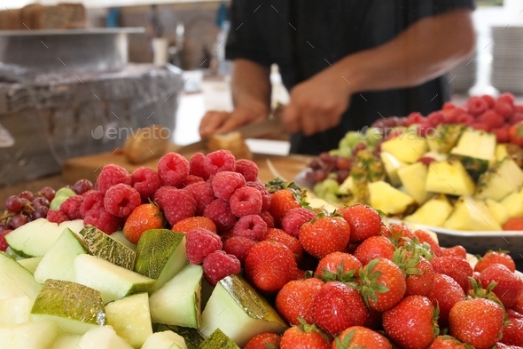 A man is slicing up bread in background, foreground with fresh strawberries, raspberries, melon,