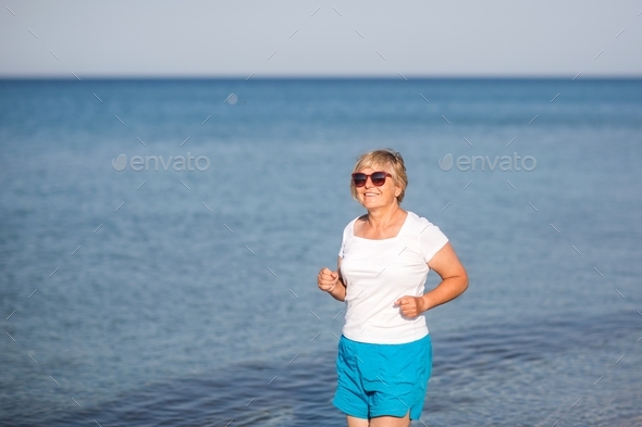 Happy senior woman runs along beach on background of sea.