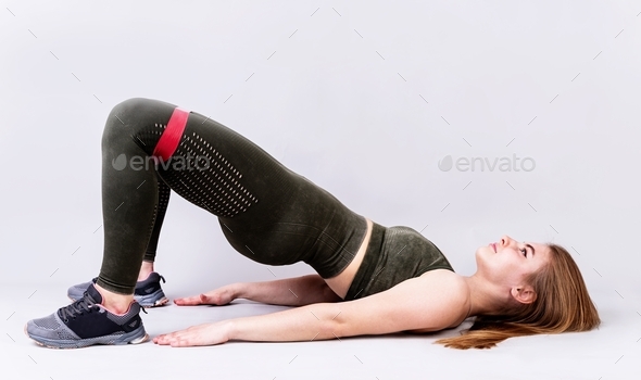 Front view of young sportswoman working out with rubber resistance bands doing hip bridge on gray
